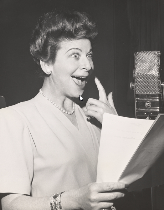 Photo of Fanny Brice recording The Baby Snooks on radio. She is holding a script, bringing a hand to her mouth and smiling widely with a childish expression of delight.