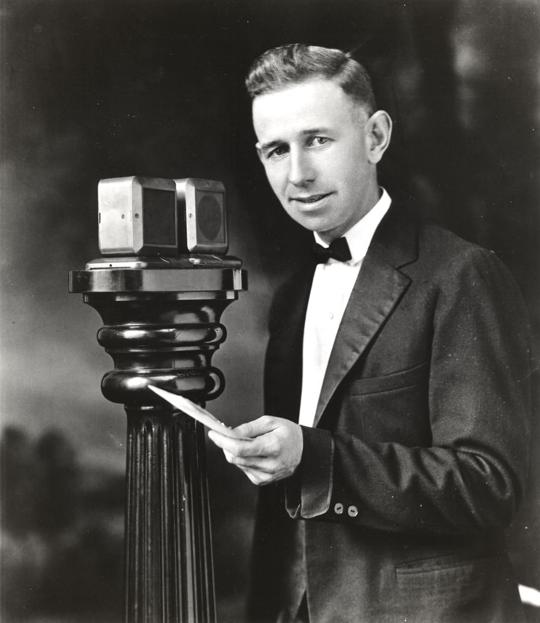 Photo of KDKA radio announcer Harold W. Arlin in 1921. He's holding a piece of paper and standing at a microphone consisting of two boxes perched on a large wooden column.