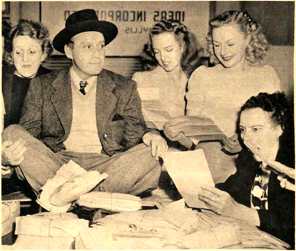 A photo of Jack Benny sitting on a table as four women look at letters submitted to the I Can't Stand Jack Benny contest in 1946