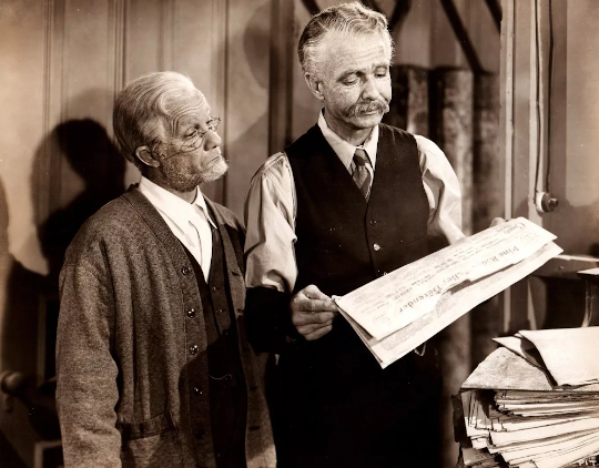 Photo from the 1946 Lum and Abner movie Partners in Time. Lum and Abner are looking at the front page of a newspaper in an office that has other newspapers stacked behind them.