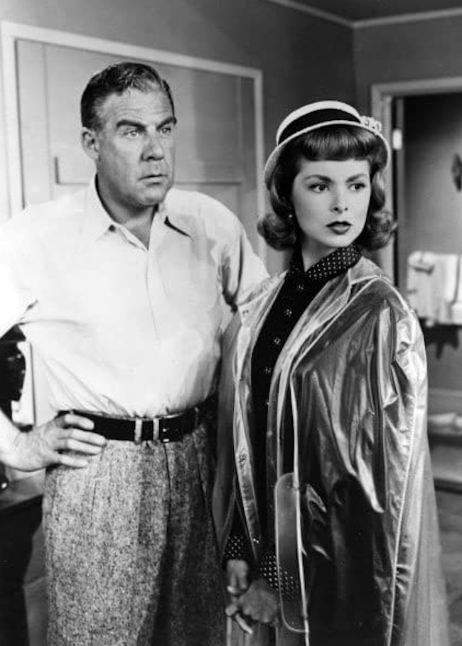 Paul Douglas and Janet Leigh in the 1951 baseball movie Angels in the Outfield. In a hotel room, Janet is wearing a coat and smart hat and has turned away from Paul with a pensive look on her face. Paul wears slacks and a white dress shirt and has his hands at his sides, looking past Janet.